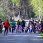 Grupa dzieci biegnąca po asfaltowej drodze w otoczeniu lasu. W tle są drzewa i dorośli kibicujący uczestnikom. Dzieci mają kolorowe ubrania i numery startowe na koszulkach.