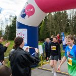 Dziewczynki w kolorowych koszulkach przechodzą przez metę biegu. Tło stanowi zielona roślinność i widownia. Nad nimi wisi kolorowy łuk z napisem.