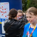 Na zdjęciu są dzieci z medalami po zakończeniu biegu. Kolorowe stroje na tle zieleni i ludzie w tle. Na obok widoczna jest mapa z kalendarzem.