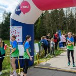 Biegaczka w niebiesko-zielonym stroju mija nadmuchaną bramę z napisem „Ale czas!”. W tle drzewa i kibice. Słoneczny dzień.