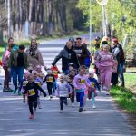 Grupa małych dzieci uczestniczy w biegu ulicznym. W tle las, dominują kolory zieleni i błękitu. Dzieci mają numery startowe.