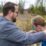 Mężczyzna wręcza medal dziewczynce na otwartej przestrzeni. W tle kilka dzieci, drzewa i trawiaste pole. Scena jest kolorowa i wiosenna.