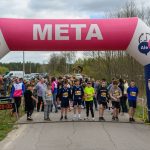 Zdjęcie przedstawia start biegu z dziecięcymi uczestnikami pod różowo-niebieskim łukiem z napisem META. W tle widać drzewa i samochody, a z boku stoi mężczyzna z mikrofonem. Na zegarze widnieje czas 0:00:00