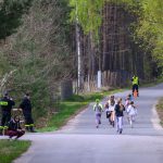 Grupa dzieci biegnie po asfaltowej drodze otoczonej zielonym lasem. Na poboczu stoi kilku strażaków w czarnych mundurach. Tło tworzą drzewa z wiosennym listowiem.