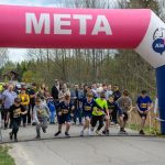Na zdjęciu grupa dzieci startuje w biegu spod różowego łuku z napisem „META”. Tło stanowi zalesiony krajobraz, a z boku stoi tłum ludzi. Widoczna jest tablica z czasem 0:00:00.