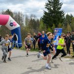 Grupa dzieci rozpoczyna bieg przy różowo-niebieskim łuku startowym. Ubrane są w sportową odzież, niektóre w jaskrawe kolory. W tle widać drzewa i ludzi kibicujących.