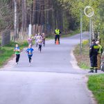 Na zdjęciu grupa dzieci biegnie po asfaltowej drodze otoczonej zielonym lasem. W tle stoi dwóch strażaków i ochroniarz. Dzieci mają kolorowe koszulki i numery startowe.