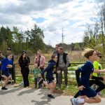 Grupa dzieci biegnie w wyścigu, na tle drzew i nieba, z kibicującymi osobami z boku drogi. Kolory to głównie zieleń, błękit i szarość. W tle widoczni widzowie z aparatami.