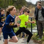 Dzieci biegną w zawodach na zewnątrz. Tło to zielony krajobraz i widzowie. Ubrani są w sportowe stroje w żywych kolorach.