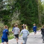 Dzieci biegają po ścieżce. Ubrane w kolorowe stroje sportowe, otoczone zielenią drzew. Droga prowadzi przez las.