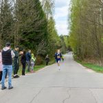 Biegacz w niebieskim stroju biegnie po asfaltowej drodze w lesie. W tle widać drzewa i dopingujących ludzi. Po lewej stronie stoi niebieski baner.