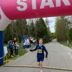 Młody biegacz w granatowym stroju przekracza linię mety, pod różowym balonowym napisem START. W tle drzewa i obserwatorzy.