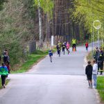 Na drodze między drzewami biegnie grupa dzieci, ubranych w kolorowe stroje sportowe. Po bokach stoją dorośli, niektórzy w ciemnych ubraniach. Tło jest zdominowane przez zieleń i wysokie drzewa.