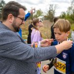 Mężczyzna wręcza medal chłopcu w koszulce z numerem 259. W tle osoby, zielone krzewy i pochmurne niebo. Atmosfera sportowej uroczystości.