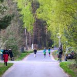 Na zdjęciu widoczni są biegnący ludzie na leśnej drodze. Tło stanowią zielone drzewa i krzewy. Wokół znajdują się osoby obserwujące oraz zaparkowany samochód.