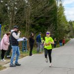 Młody biegacz w jasnozielonej koszulce biegnie, wokół kibice i las. Mężczyzna z mikrofonem komentuje. Jest jasne, pochmurne niebo.