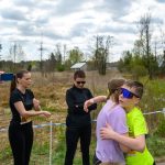 Dwoje dzieci w kolorowych koszulkach przytula się na tle natury. Widać niebieski balon z mapą i dorosłych obserwujących sytuację. Pogoda jest pochmurna, a w tle zieleń i budynki.