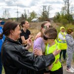 Grupa ludzi na zewnątrz, w tym dzieci w sportowych okularach, ściska się. Tło to teren z drzewami i niebem z chmurami. Dorośli robią zdjęcia.