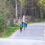 Na zdjęciu widoczne są cztery osoby biegnące drogą, ubrane w niebiesko-zielone koszulki. Po lewej stronie znajduje się zalesiony teren z zielonymi drzewami. Tło stanowi las z wysokimi drzewami i ogrodzenie wzdłuż drogi.
