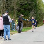Na zdjęciu młody biegacz w niebieskim stroju z numerem 264 biegnie po asfaltowej drodze. Tło stanowi zielony las i widzowie z boku. Obok drogi widoczny jest niebieski baner reklamowy.