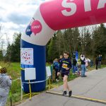 Na zdjęciu młody biegacz przekracza linię startu-meta pod różowo-niebieskim łukiem. W tle widać zielone drzewa i niebo pełne chmur. Obok kibice i obsługa zawodów dopingują biegaczy.