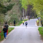 Biegacze na asfaltowej drodze pośród zielonych drzew. W tle widać zarośla i strażaków w odblaskowych kamizelkach. Jasny dzień, spokojne otoczenie.