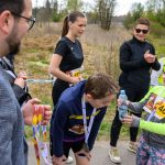 Na zdjęciu grupa ludzi po biegach, dziecko w zielonej czapce podaje wodę. Tło to sucha roślinność, część osób nosi medale. Kolory są jasne, dominują zielenie i żółcie.