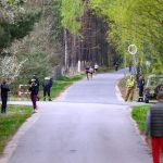 Widać asfaltową drogę przez las, ludzie biegną i spacerują. Po bokach dużo zieleni i drzewa. Ochrona i samochód po prawej.