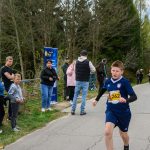 Na zdjęciu chłopiec w niebieskim stroju biegnie po asfalcie. Obok stoi grupa kibiców przy ogrodzeniu. W tle widać drzewa i niebo.