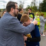 Grupa ludzi na zewnątrz, część w sportowych ubraniach. Mężczyzna w marynarce przytula chłopca w koszulce z numerem. W tle drzewa i niebo.