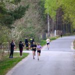 Na zdjęciu widać biegaczy na wąskiej asfaltowej drodze w lesie. Tło jest zielone, z drzewami i obserwatorami na poboczu. Kierują się do mety, co sugerują liczby na ich koszulkach.