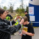 Kobieta zakłada medal dziecku z numerem 267. W tle niebieski balon i kilka osób. Słoneczny dzień na zewnątrz.