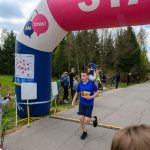 Na zdjęciu widoczny jest biegacz w niebieskiej koszulce, przekraczający linię mety z numerem startowym 263. Tło stanowi zalesiona sceneria z widocznym niebem i publicznością. Nad linią mety wisi różowy i niebieski balon z nap