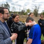 Na zdjęciu kobieta zakłada medal chłopcu w niebieskiej koszulce. W tle ludzie na tle zieleni i lekko zachmurzone niebo. Atmosfera jest radosna i uroczysta.