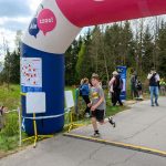 Chłopiec w szarej koszulce biegnie pod nadmuchiwanym łukiem z napisem. W tle drzewa i kibice obserwujący bieg. Pogoda pochmurna.