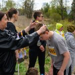 Osoba w czarnej kurtce wręcza medal chłopcu z numerem 265. W tle są drzewa i kilka innych osób. Chłopiec jest ubrany w szarą koszulkę.