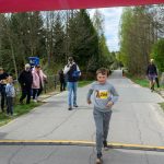 Chłopiec w szarym dresie biegnie pod kolorowym łukiem z numerem 266. W tle drzewa, osoby i niebo. Zawody na ścieżce.