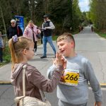 Na zdjęciu dwoje dzieci przybija piątkę na asfaltowej drodze, chłopiec w szarym stroju ma numer 266. W tle widać ludzi, drzewa i baner, dominują kolory zielony i szary. To scena z wydarzenia sportowego na świeżym powiet