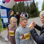 Na zdjęciu dzieci na zawodach biegowych, przyjmujące medal od uśmiechniętej osoby. W tle nadmuchiwany łuk i zalesiona sceneria. Kolory są jasne i wiosenne, dominują zielony i niebieski.