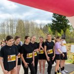 Grupa młodych osób w czarnych strojach sportowych stoi pod kolorowym łukiem startowym. W tle drzewa i niebo zasłane chmurami. Na numerach startowych widoczne numery i logo sponsorów.