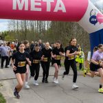 Grupa młodych kobiet biegnie w zawodach pod różowo-białym balonem z napisem META. W tle las i kibice. Start wyścigu oznaczony zegarem.