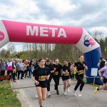 Na zdjęciu widać grupę biegaczek przekraczających linię mety pod różowo-białym łukiem z napisem META. Tło stanowi zielony krajobraz z drzewami i tłumem widzów. Ubrania biegaczek są głównie czarne, z