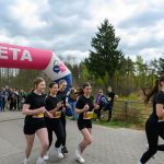Cztery biegaczki w czarnych strojach przekraczają różową linię mety. W tle widoczna jest zielona roślinność i kibice. Nad nimi niebo jest pochmurne.