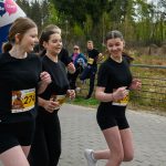 Trzy młode biegaczki w czarnych strojach uczestniczą w wyścigu. W tle widać balon startowy i zieleń drzew. Po prawej stronie stoi niebieski baner reklamowy.