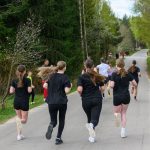 Grupa ludzi biegnie po asfaltowej drodze. Wokół widoczne są drzewa i zieleń, tworzące naturalne tło. Większość uczestników ubrana jest na czarno.