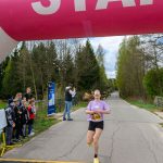 Biegaczka przekracza linię mety pod różowym, nadmuchiwanym łukiem z napisem START. W tle drzewa i kibice. Dzień jest pochmurny.