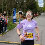 Biegaczka w fioletowej koszulce z numerem 269 biegnie drogą. Ma wielokolorowe okulary. W tle las i kibicująca grupa ludzi.