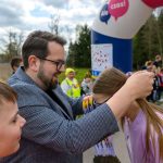 Mężczyzna w okularach zakłada medale dziewczynie w fioletowej bluzie. Jasne, zachmurzone niebo i zielone tło. W tle grupa ludzi.