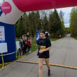 Uczestniczka biegu w czarnym stroju mija metę pod kolorowym, nadmuchiwanym łukiem. W tle widać zielone drzewa i widzów przy trasie. Pogoda jest pochmurna.