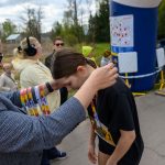 Mężczyzna wręcza medal dziewczynie na mecie biegu. W tle drzewa i balon reklamowy. Kilka osób obserwuje z boku.